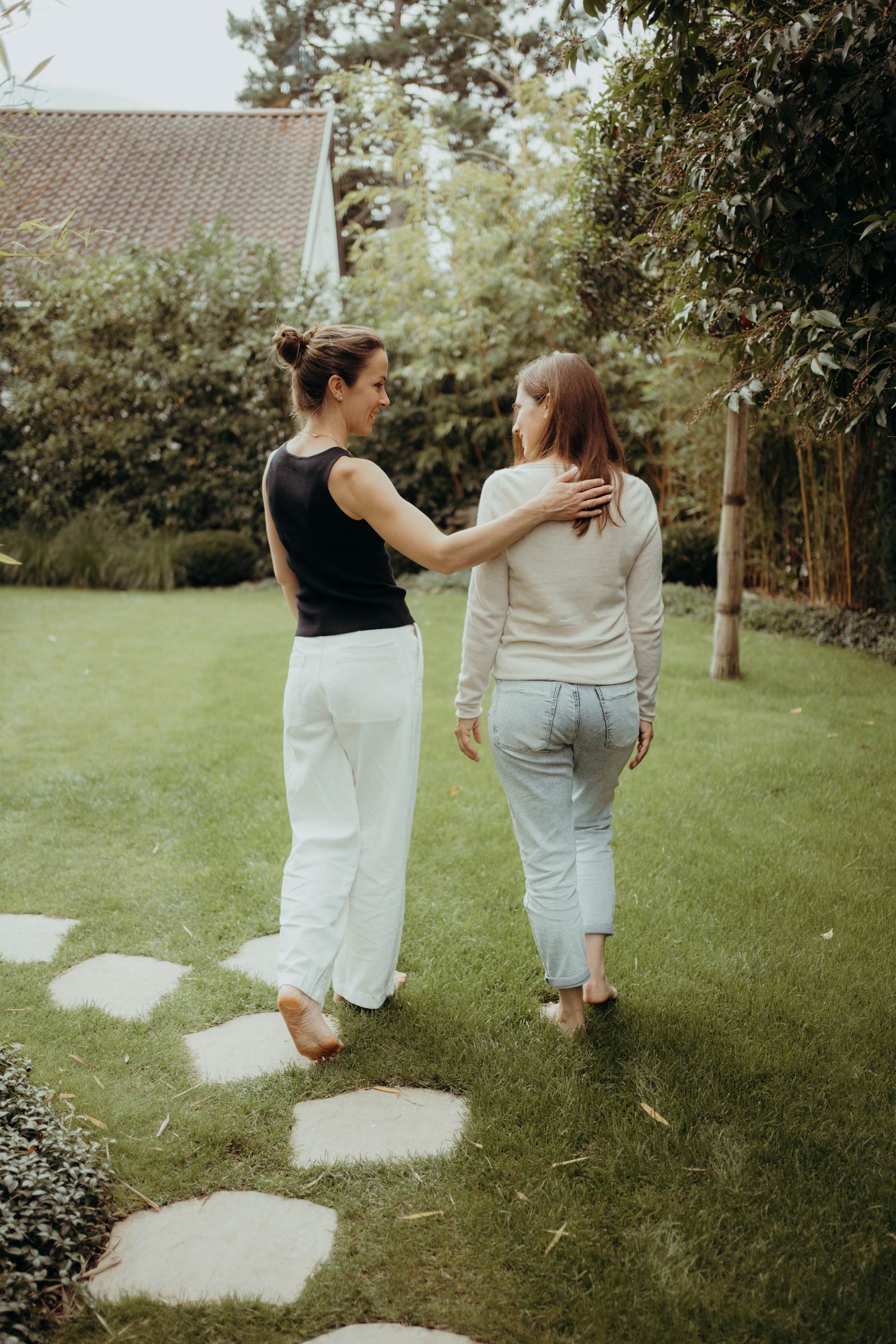 Zwei Frauen gehen nebeneinander über eine Wiese mit Trittsteinen, eine legt der anderen den Arm um die Schulter.